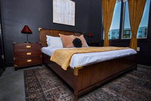 Habitación de hotel con cama de madera, mesita de noche, lámpara, alfombra estampada y ventana con cortinas.