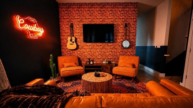Ein gemütliches Wohnzimmer mit Backsteinwand, zwei orangefarbenen Sesseln, einem Ledersofa, einem runden Couchtisch, einer Gitarre, einer Banjo und einem Neonlichtschild mit der Aufschrift 'The Cowboy Club'.
