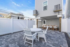 Une terrasse avec une table ronde et des chaises, une clôture blanche et un bâtiment équipé d’un climatiseur.