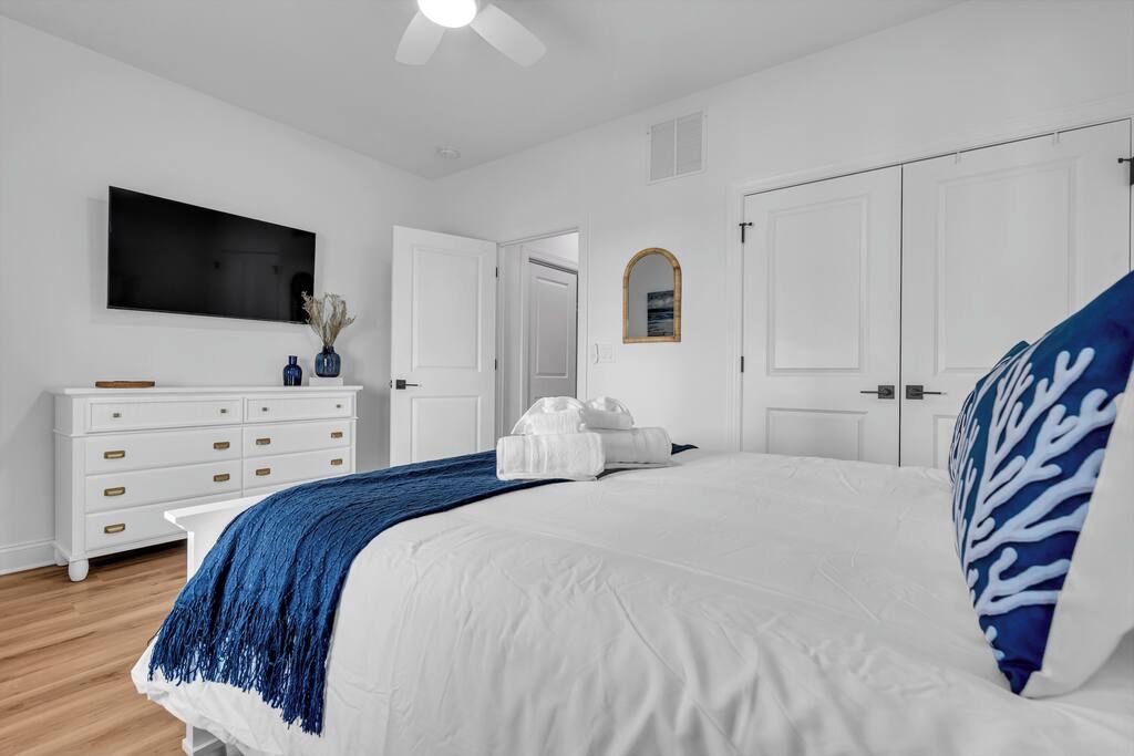A bedroom with a large bed, white walls, a ceiling fan, a flat-screen TV, a dresser, and a mirror.