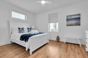 Une chambre avec un lit blanc, un plancher en bois, un ventilateur de plafond, une fenêtre avec des stores, un tableau au mur et une petite table blanche.