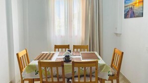 Un comedor con una mesa dispuesta para cuatro, sillas de madera, una ventana con cortinas translúcidas y un cuadro en la pared.