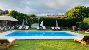 Piscina con sillones de descanso y sombrillas, rodeada de un césped cuidado y una casa al fondo.