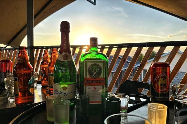 A bar area with various bottles and glasses, a wooden railing, and a view of the sunset.