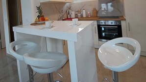 A modern kitchen with white cabinets, a stainless steel range hood, and a built-in oven. There are two white bar stools and a small island with a white countertop. - Beachfront Apartment With Balcony and Sea View (Vrsi)