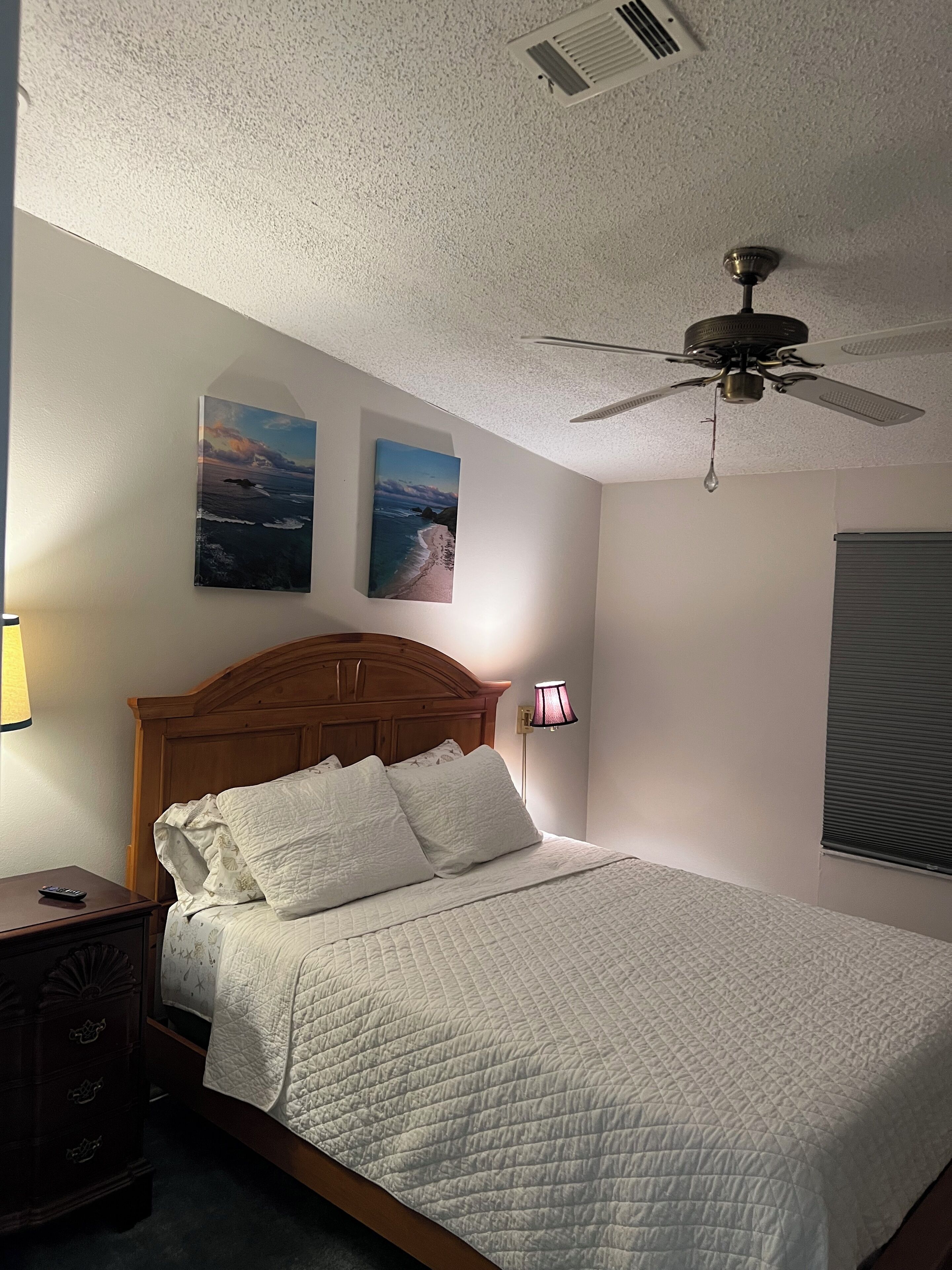 Un dormitorio con cabecera de madera, cama con ropa blanca, dos cuadros colgados en la pared, un ventilador de techo, una lámpara de noche y una mesita de noche con lámpara.