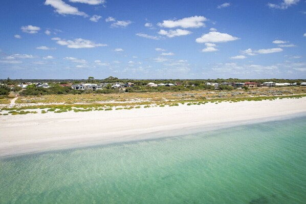 A coastal landscape with a sandy beach, clear turquoise waters, and a row of houses in the background.