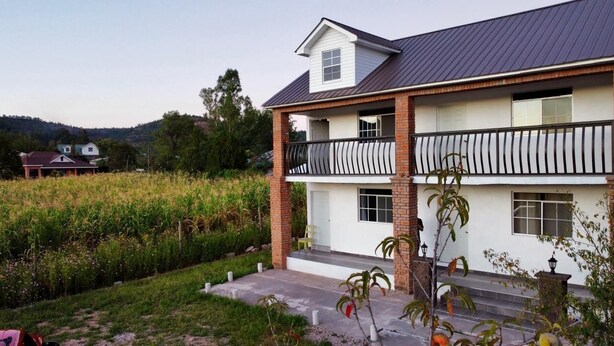 A two-story house with a balcony, surrounded by a garden and a field.