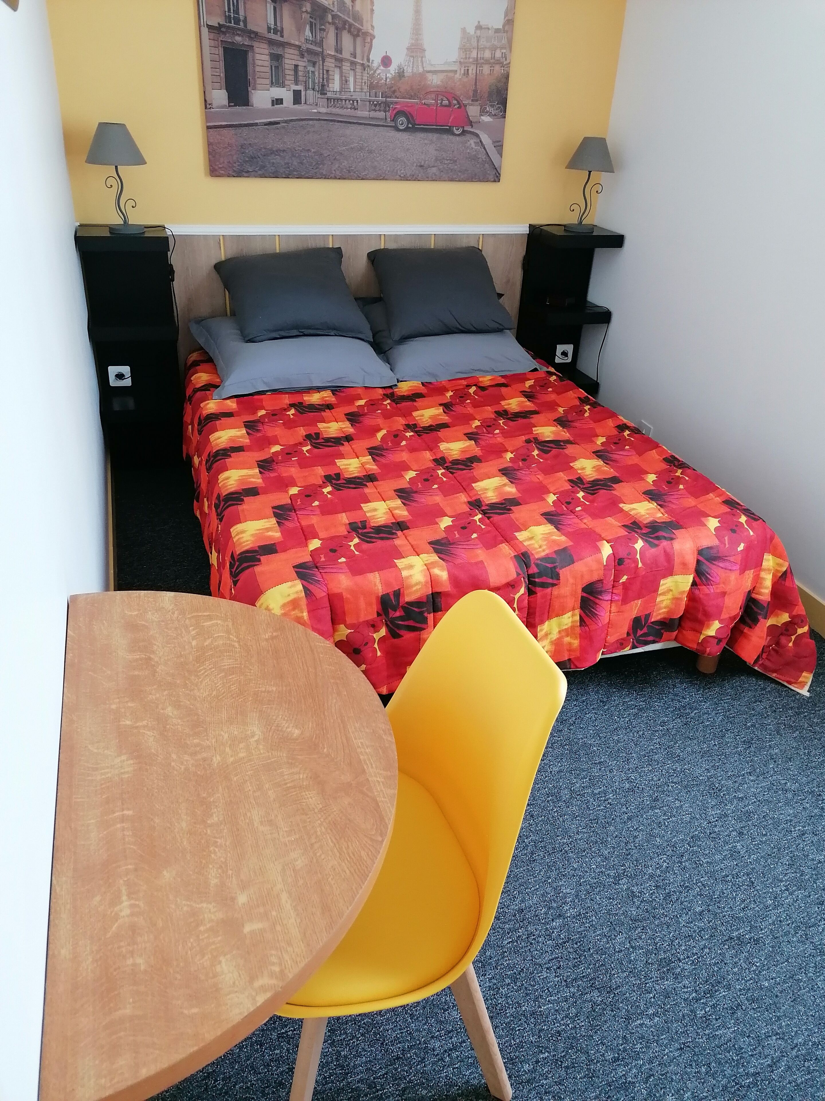 Une chambre à coucher avec un lit, deux tables de chevet avec des lampes, une table en bois, une chaise jaune et un tableau représentant un paysage urbain accroché au mur.