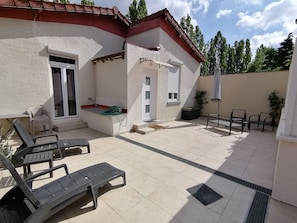 Eine Terrasse mit weißer Fassade, einer Schiebetür und einem kleinen Fenster. Zwei Lounge-Stühle und ein Tisch mit Sitzgelegenheiten sind vorhanden.