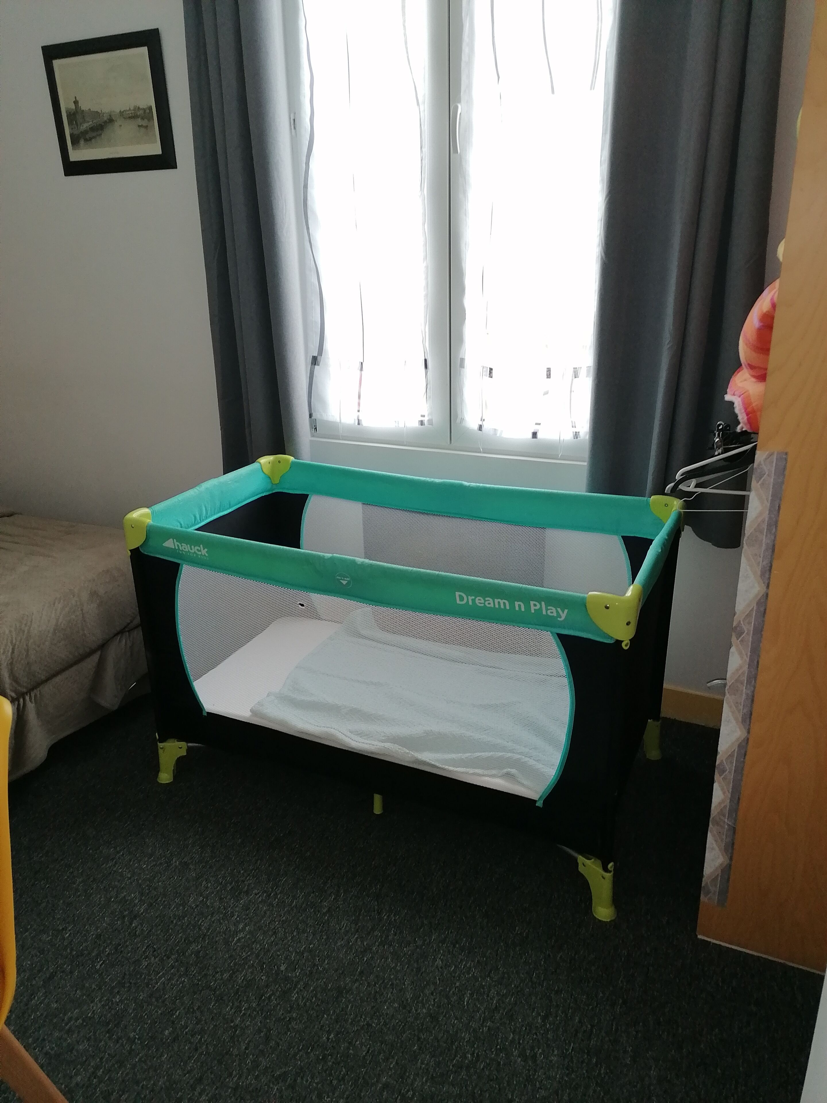Une pièce avec un berceau, une fenêtre avec des rideaux, un tableau encadré au mur et un lit.