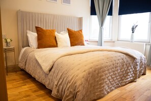 Una cama bien hecha con edredón color beige y dos almohadas marrones, una mesita de noche con un jarrón de flores y una ventana con cortinas.