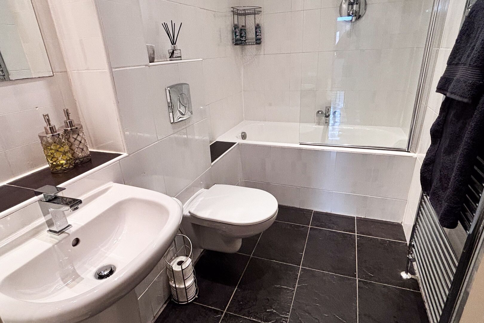 A modern bathroom with a white sink, toilet, and bathtub. There are toiletries on the counter and a towel rack.