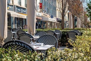 Outdoor dining tables with chairs, a building with signage, and a hedge in the foreground. - Mobility lease l Equipped & Paris 10 mins away (Neuilly-sur-Seine)