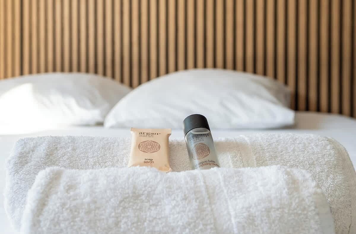 Een bed met witte kussens, een witte handdoek, een fles shampoo en een zeepstuk op het bed.
