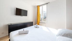 A hotel room with a bed, a flat-screen TV mounted on the wall, a radiator, and a window with yellow curtains.