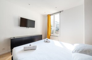 A hotel room with a bed, a flat-screen TV mounted on the wall, a radiator, and a window with yellow curtains.