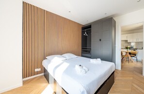 A modern bedroom with a bed, wooden panel wall, and a kitchenette in the background.