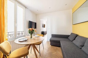 A modern living room with a sofa, a dining table with chairs, a TV mounted on the wall, and a balcony with a railing.