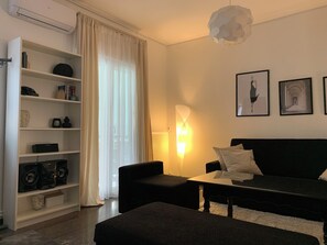 A modern living room with a black sofa, a white coffee table, and a black armchair. There is a white bookshelf with various items on the shelves. A large window with sheer curtains allows natural light into the room. - Your home in Athens (Galatsi)