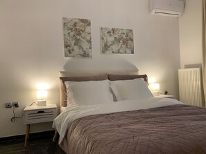 A neatly made bed with a brown quilt, two wall-mounted artworks, a bedside lamp, and a white bedside table with a drawer. - Your home in Athens (Galatsi)