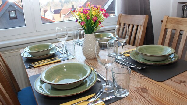 Une table à manger dressée avec de l’assiette verte, des bols, des couverts et un vase rempli de fleurs.