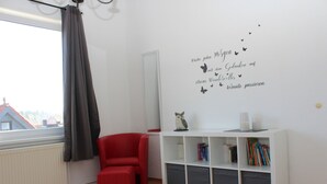 A room with a red chair, a white bookshelf, and a window with a view of buildings. - Hexenquartier apartment in a central location in Braunlage (Braunlage)