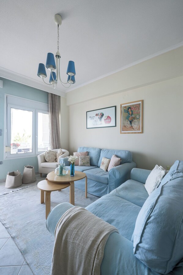 Una sala de estar con un sofá azul claro, una mesa de centro redonda de madera y una araña. Hay dos cuadros enmarcados en la pared y una ventana con cortinas.