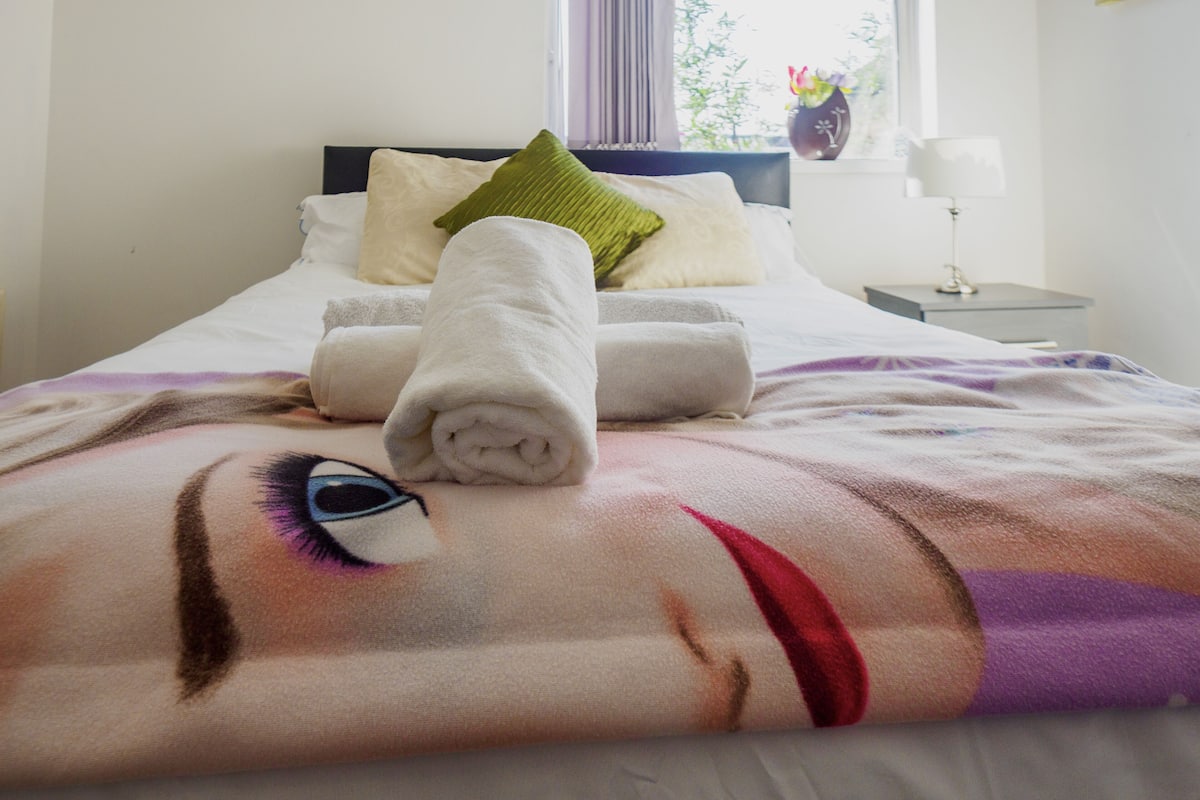 A neatly made bed with a rolled towel, pillows, and a blanket with a face design.