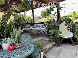 Ein überdachter Patio-Bereich mit einem Korbstuhl, einem kleinen Tisch und verschiedenen Blumentöpfen.