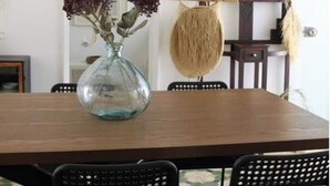 Un comedor con una mesa de madera, sillas negras, un florero de vidrio con flores y un tapiz de macramé en la pared.