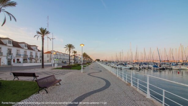 Un paseo marítimo con un banco, una hilera de edificios y un puerto deportivo con barcos.