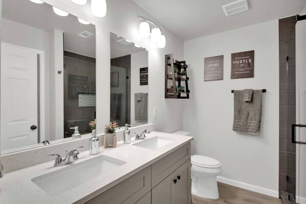 A modern bathroom with a large mirror, two sinks, a toilet, and a shower area.