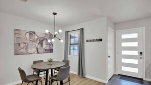 Un coin repas comprenant une table ronde et quatre chaises, un luminaire suspendu, une fenêtre avec des stores et un porte-manteau.