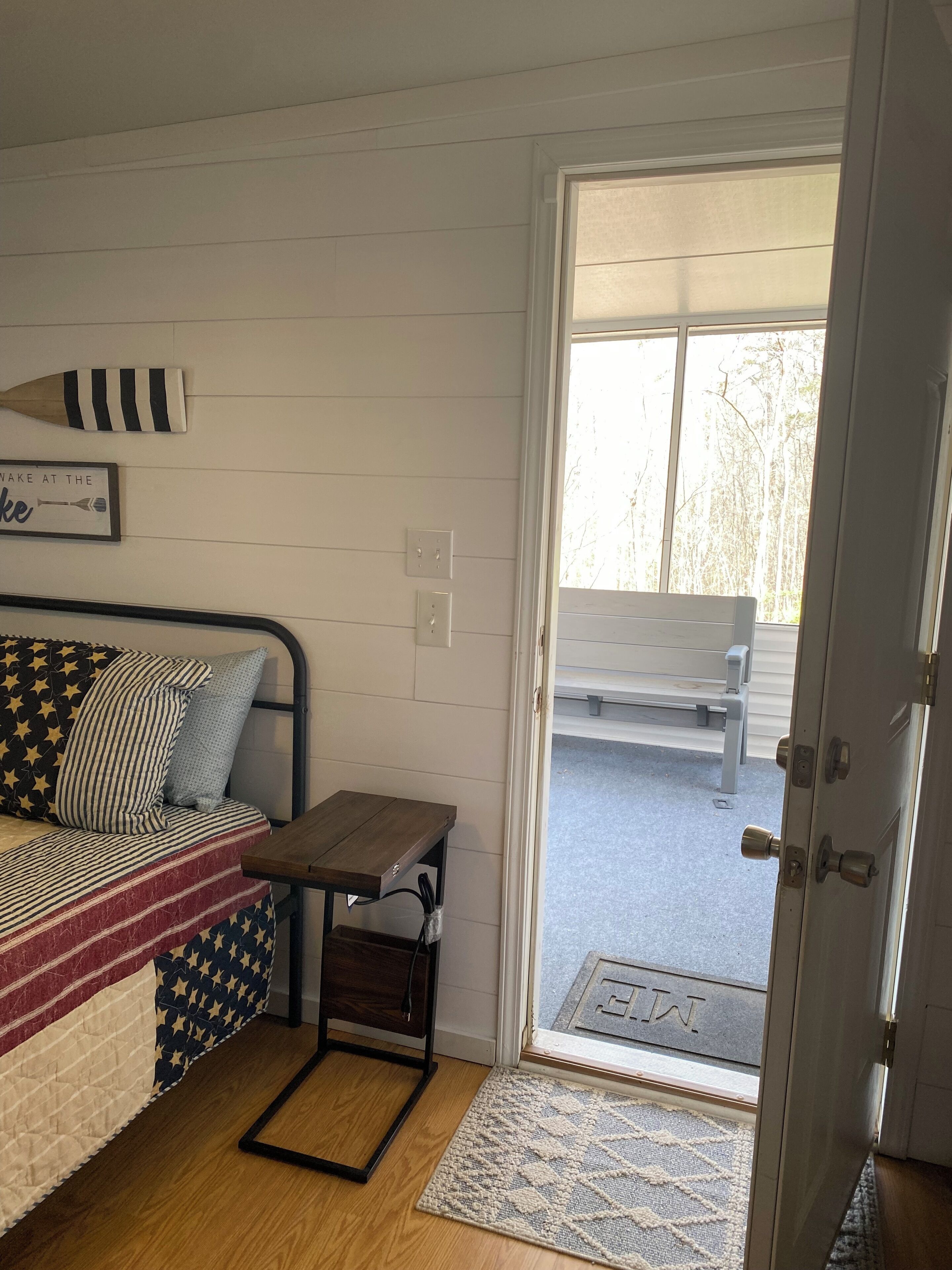 A room with a bed, a small table, a bench outside, and a door with a doormat.