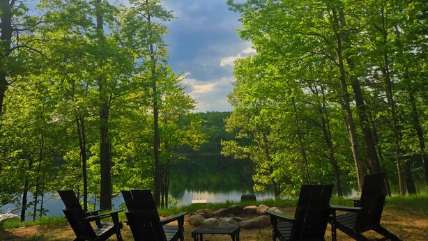 A lakeside campsite with four Adirondack chairs and a small table, surrounded by tall trees and a clear sky. - Fall is beautiful on Mystery Lake! Book now for fishing, campfires and boating! (Spooner)