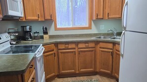 A kitchen with wooden cabinets, a refrigerator, a stove, and a window with a view of trees. - Fall is beautiful on Mystery Lake! Book now for fishing, campfires and boating! (Spooner)