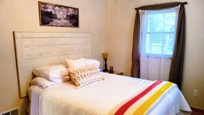 A neatly made bed with a white quilt and a colorful striped blanket, a wooden headboard, a framed picture on the wall, and a window with curtains. - Fall is beautiful on Mystery Lake! Book now for fishing, campfires and boating! (Spooner)