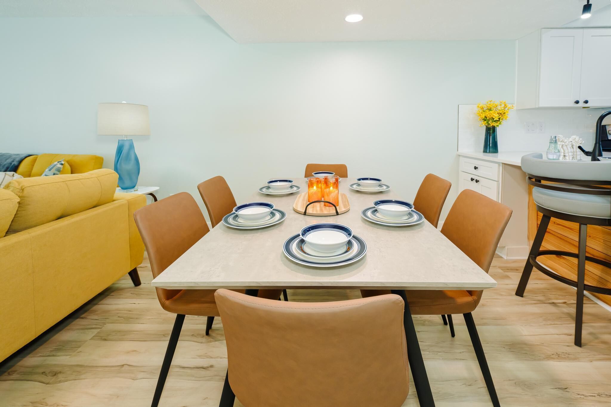 Un comedor con una mesa dispuesta para cuatro personas, rodeada de sillas, en una habitación con un sofá amarillo y una lámpara.