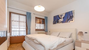 A bedroom with a bed, a nightstand, a window with curtains, a radiator, and a painting on the wall. - Appartamento Iori (Canazei)
