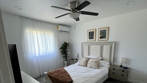 Un dormitorio con cama, mesitas de noche, ventilador de techo, televisor y ventana con cortinas.