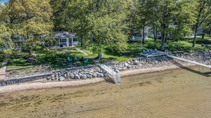 A riverside property with a deck, outdoor seating, and a stone retaining wall. - Lakefront 3-bedroom cottage on Sand Point with AC, WiFi, King bed, Firepit (Pigeon)