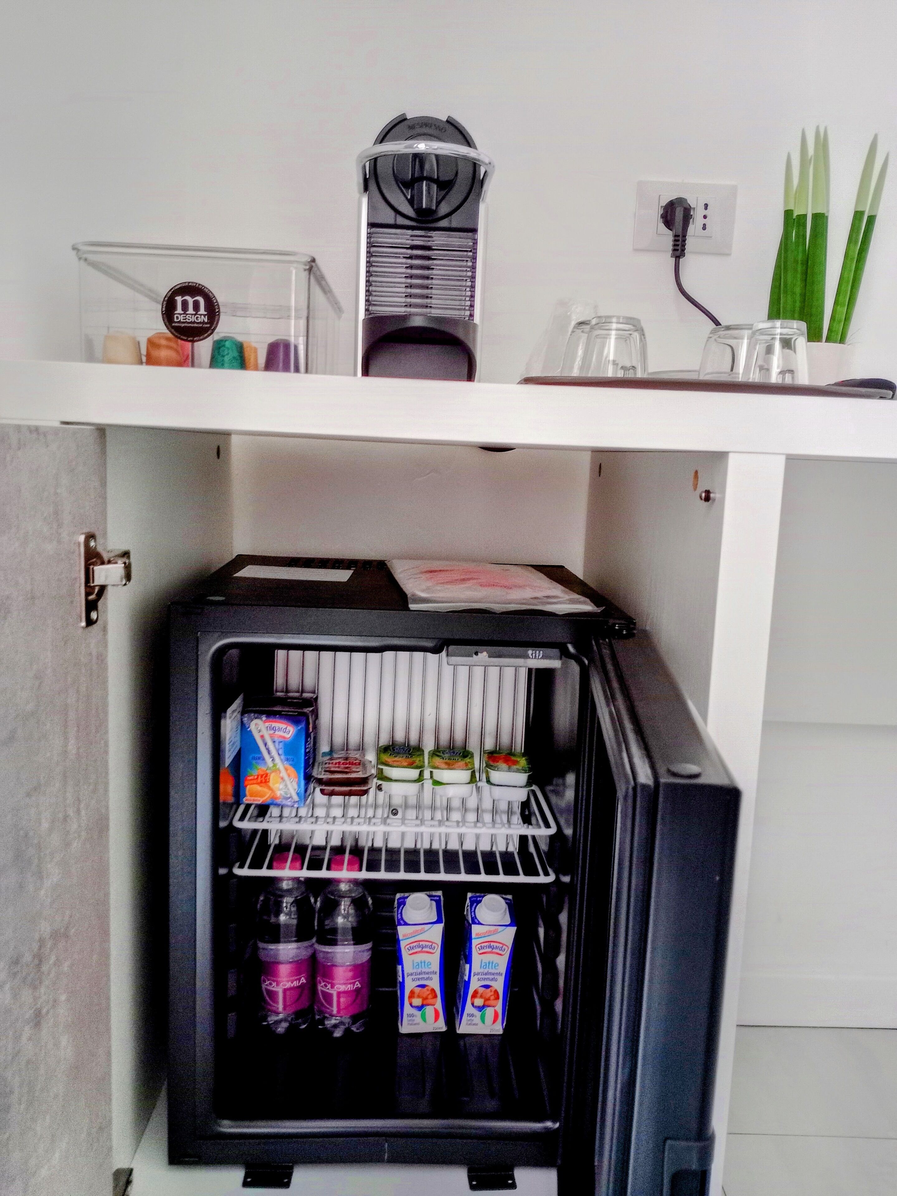Un refrigerador compacto con dispensador de leche y jugo, una cafetera y un pequeño estante con objetos decorativos.