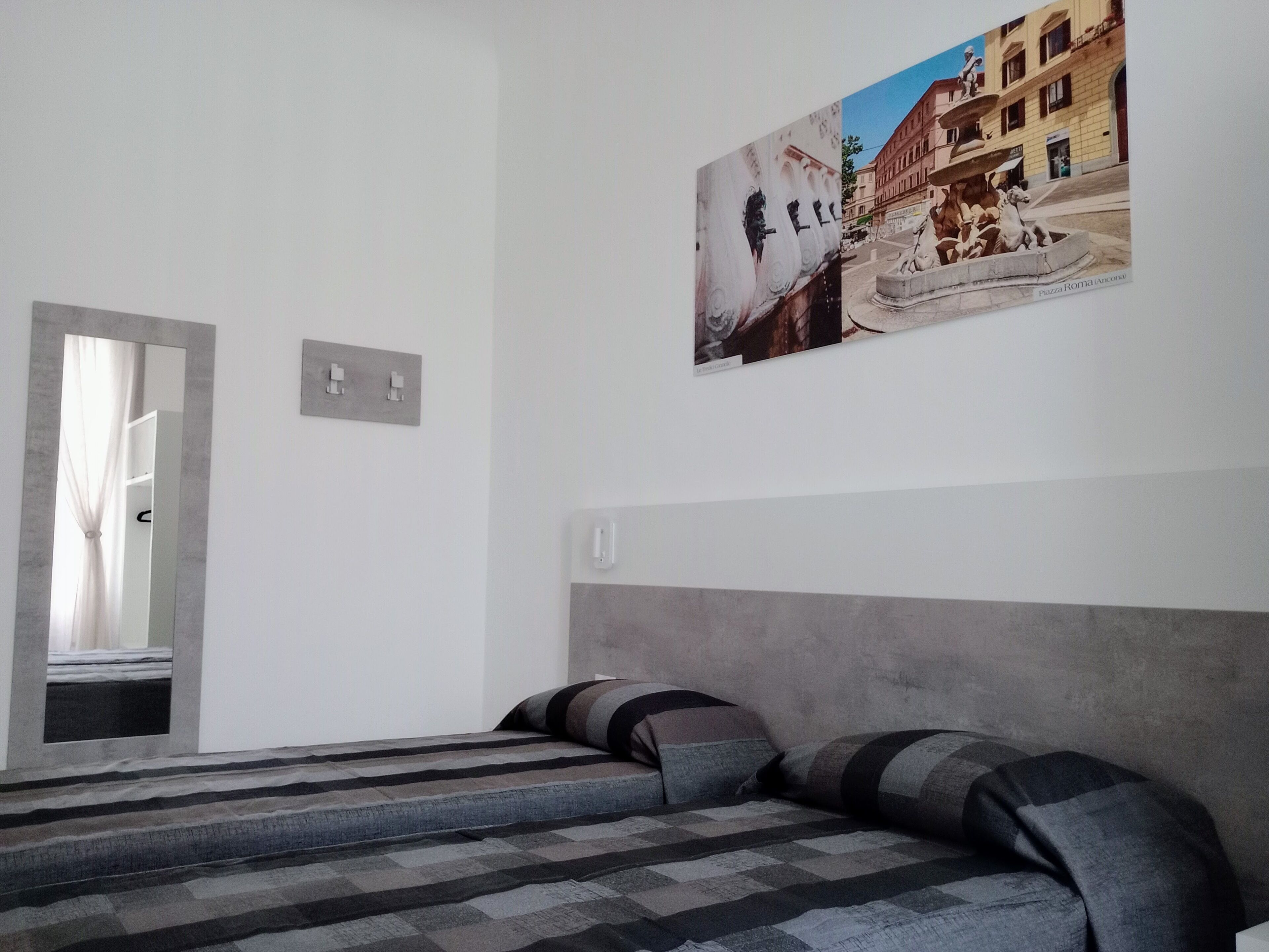 A bedroom with a bed, a mirror, a wall-mounted light switch, and a framed picture of a building.