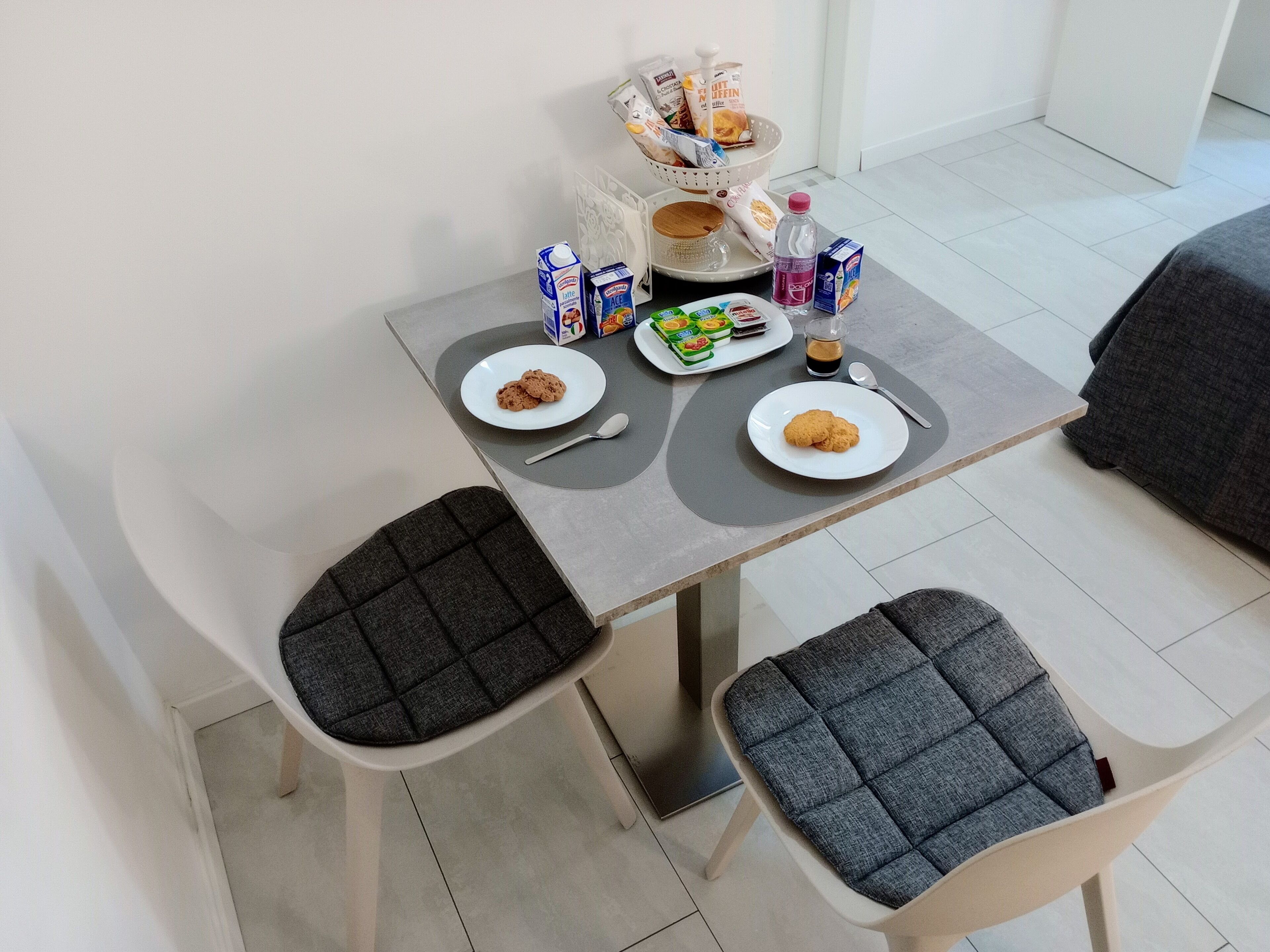 Un pequeño comedor con dos sillas grises acolchadas, una mesa con platos de comida, un cesto de pan y una botella de agua.