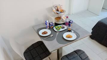 Un pequeño comedor con dos sillas grises acolchadas, una mesa con platos de comida, un cesto de pan y una botella de agua.