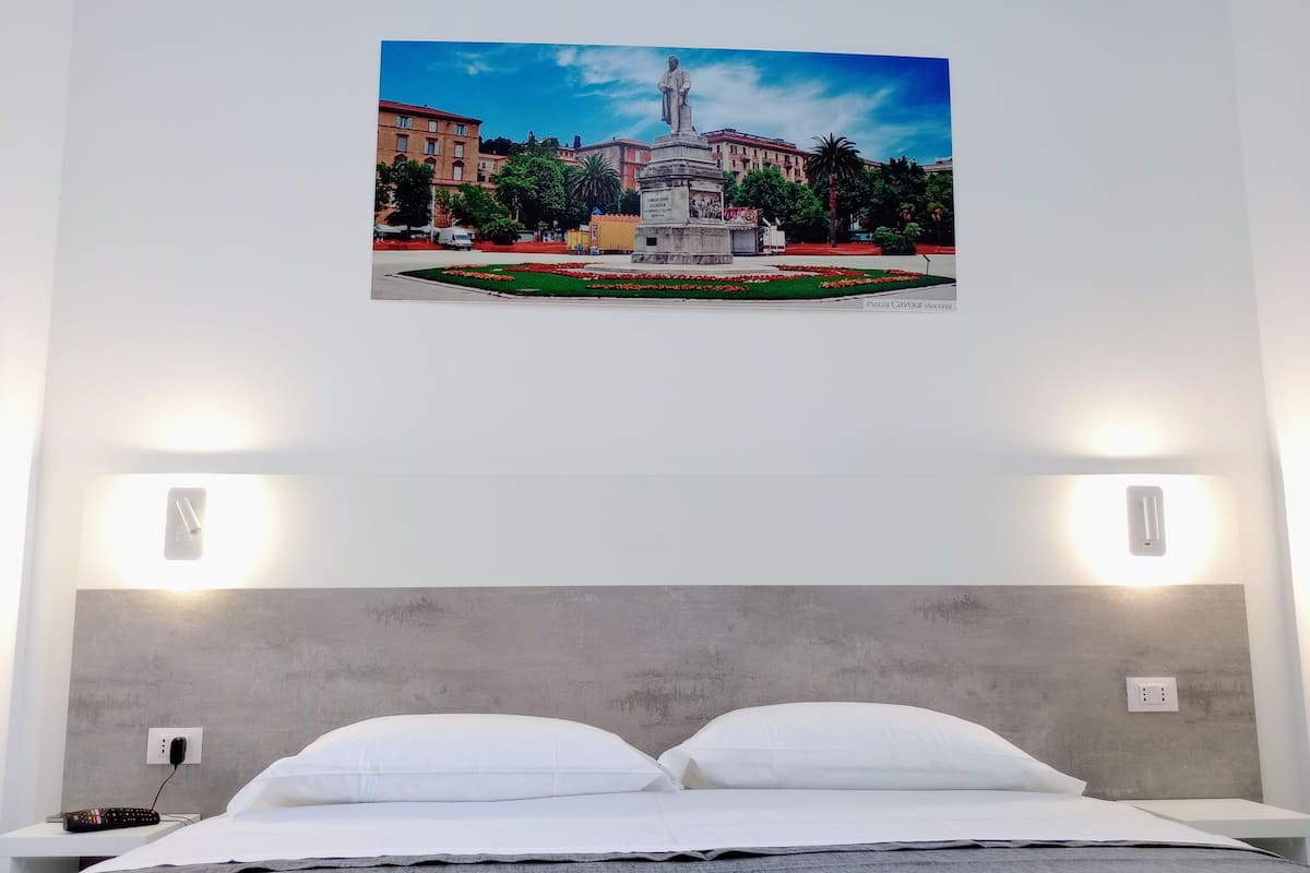 Quarto de hotel com cama, almofadas brancas, cabeceira cinzenta e quadro na parede mostrando uma praça com uma estátua.