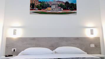 Una camera d'albergo con un letto, cuscini bianchi, una testiera grigia e un quadro appeso alla parete raffigurante una piazza con una statua.
