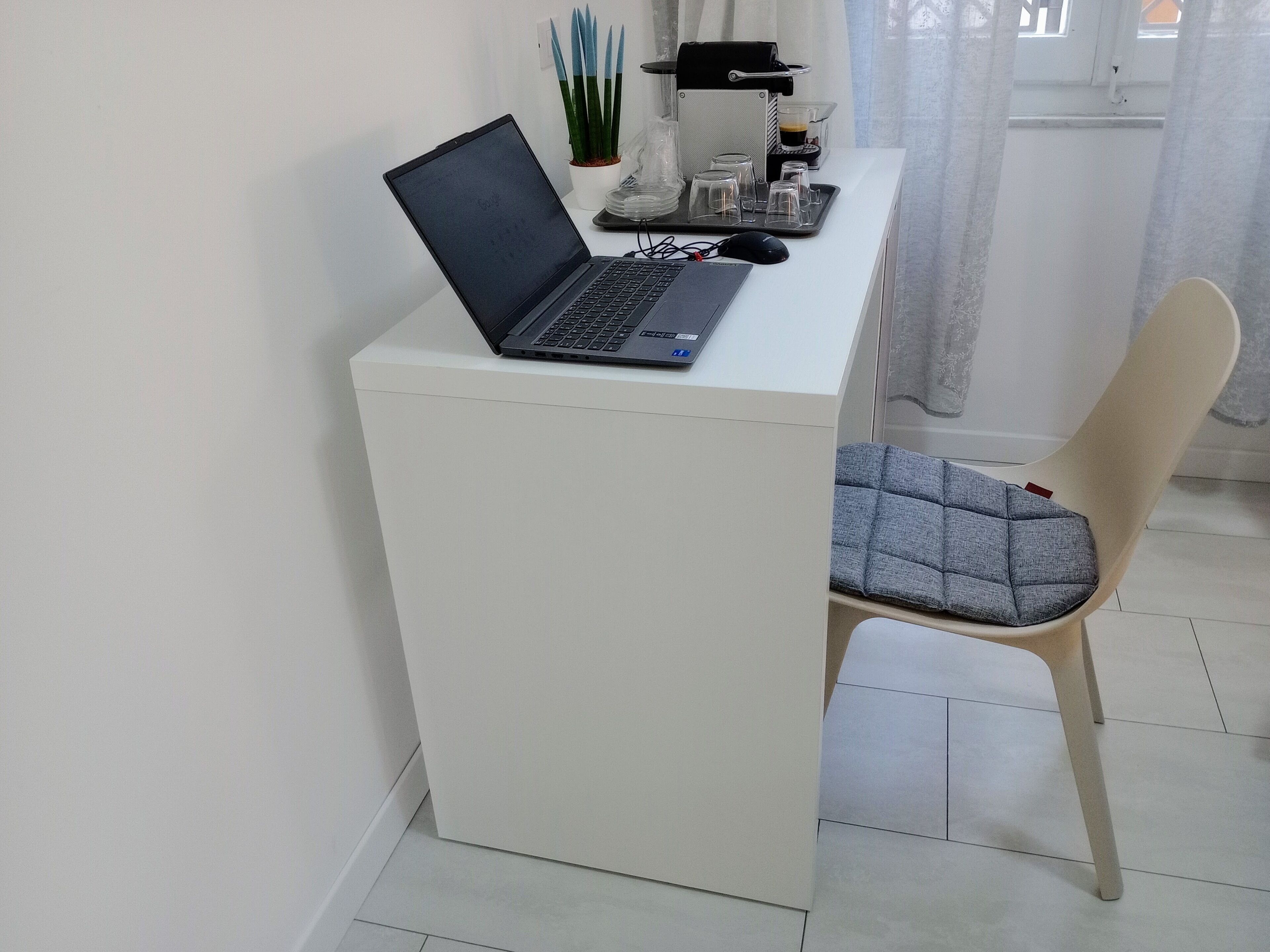 A laptop on a white desk with a coffee machine, a glass of coffee, and a potted plant.