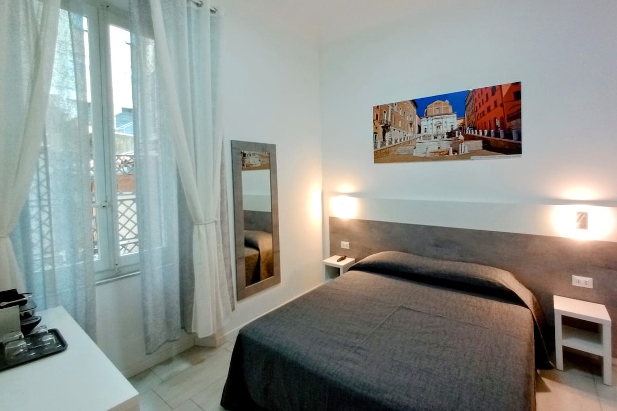 Quarto de hotel com cama, mesa de cabeceira, espelho e janela com cortinas.